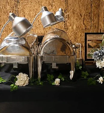 Covered chafing dishes under heat lamps with a Ponnus Catering sign at an event